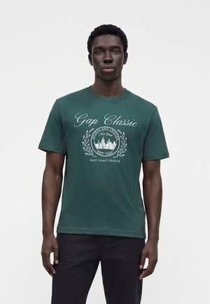 Homme portant un t-shirt Gap Classic vert foncé avec un texte graphique blanc et un motif de paysage urbain, associé à un pantalon noir, debout devant un fond blanc.