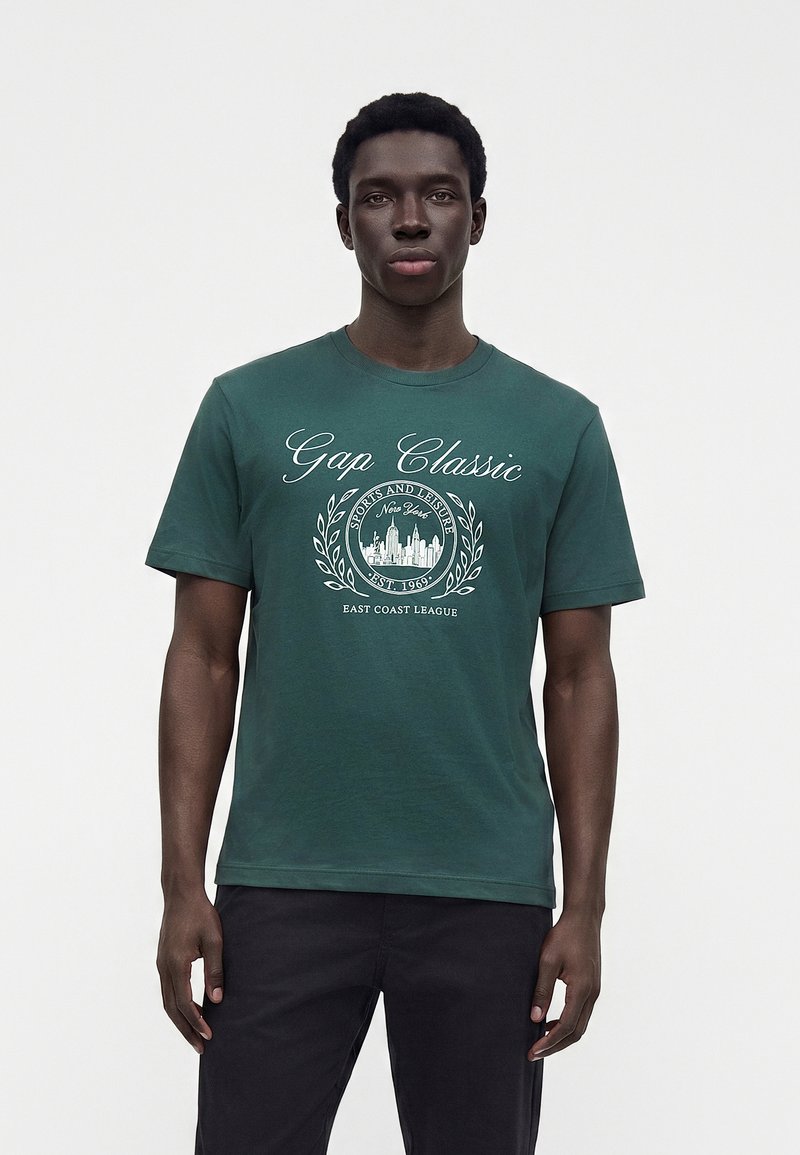 Άνδρας που φοράει σκούρο πράσινο κλασικό t-shirt της Gap με λευκό γραφικό κείμενο και σχέδιο αστικού τοπίου, συνδυασμένο με μαύρο παντελόνι, που στέκεται μπροστά σε λευκό φόντο.