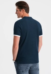 Námorně modré polo tričko s bílými akcenty na límci, vyrobené z texturované tkaniny. Krátké rukávy a boční rozparky na lemu. Model zobrazen zezadu.