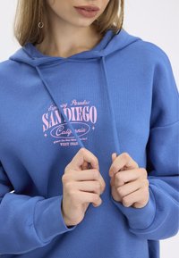 Sudadera con capucha de tela azul brillante con un estampado gráfico rosa centrado en el pecho que incluye texto. Los puños y el dobladillo son de canalé.