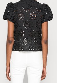 Blouse noire à manches courtes avec broderie florale en œillets et manches bouffantes, portée avec un pantalon blanc taille haute, vue de dos.