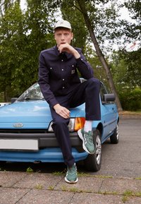 Navy work shirt and pants with pockets, light-colored baseball cap, and green sneakers. Posed on a blue car with a textured surface.