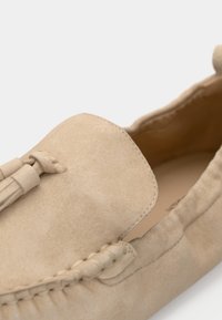 Mocassins en daim beige avec une texture lisse, un bout arrondi et un détail décoratif à pampilles sur le devant. Couture le long du bord.