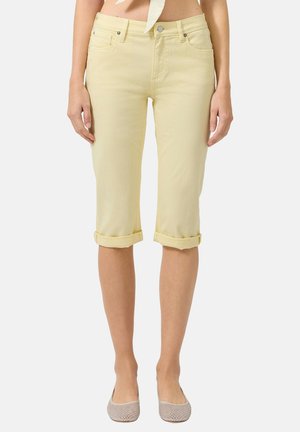 Vrouw die lichtgele capri-broeken met opgerolde boorden en lichtbeige instapschoenen draagt, staand met ontspannen armen langs het lichaam.