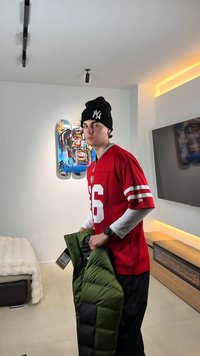Jonge man in rood sportshirt en zwarte muts die een groene donzen jas vasthoudt in een moderne kamer met skateboardmuurkunst en elektrische open haard.