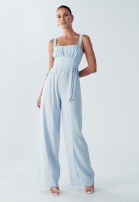 Jasnoniebieski kombinezon z kwadratowym dekoltem, szerokimi nogawkami i wiązaniem w talii. Wykonany z miękkiego materiału, z regulowanymi ramiączkami.