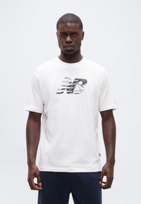 Άνδρας όρθιος που φοράει λευκό μπλουζάκι με ασπρόμαυρο στυλιζαρισμένο λογότυπο "NB" και σκούρο ναυτικό μπ λε παντελόνι πάνω σε απλό ανοιχτό γκρι φόντο.