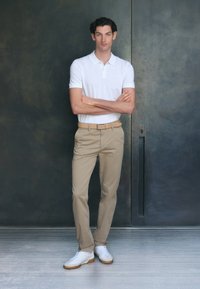 Camisa polo blanca de manga corta, chinos beige y zapatillas blancas. El modelo está de pie contra un fondo oscuro y texturizado, con los brazos cruzados.