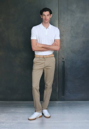 White polo shirt with short sleeves, beige chinos, and white sneakers. Model stands against a dark textured background, arms crossed.