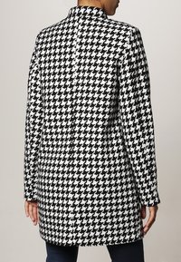 Svart och vit houndstooth-kappa med krage och långa ärmar, med en ren bakseam och mellanhög design.