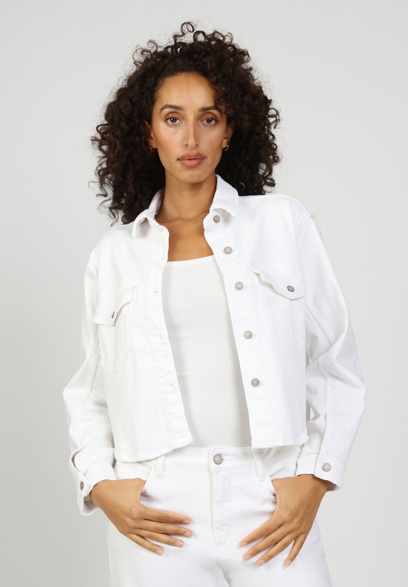 White cropped denim jacket featuring a classic collar, flap chest pockets, and snap buttons. Worn over a white tank top and matching pants.