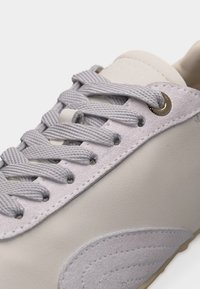 Κοντινή λήψη μπεζ αθλητικού παπουτσιού με γκρι χοντρές κορδόνια περασμένα μέσα από μεταλλικές οπές και σουέντ λεπτομέρειες κοντά στην σόλα.