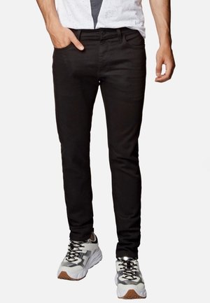 Man wearing slim-fit black jeans, white and gray sneakers, with left hand in pocket and right hand relaxed at side.