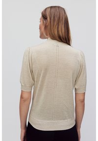 Beige kurzärmliger Pullover mit subtiler Ausstrahlung, hohem Kragen, Knopfdetails im Rücken und einer vertikalen Reihe von kleinen Öffnungen.