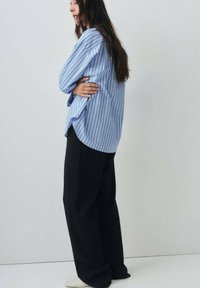 Camicia oversize a strisce blu e bianche con la parte posteriore lunga, abbinata a pantaloni neri larghi e scarpe di colore chiaro.