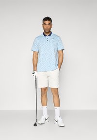 Light blue polo shirt with a navy collar and small black pattern, paired with white shorts, white socks, and grey-accented white shoes. Holding a golf club.
