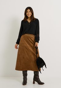 Top negro de manga larga con detalles de encaje, falda maxi de ante marrón, cinturón de cadena dorada, bolso de flecos negro y botas de tacón altas marrones.