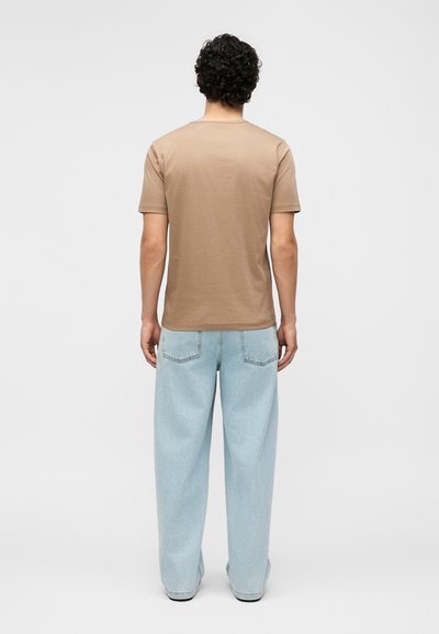 Homme aux cheveux bouclés portant une chemise beige à manches courtes et un jean bleu clair ample, debout de dos sur un fond uni.
