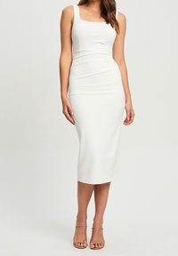 Bílé midi šaty na tělo se čtvercovým výstřihem, širokými ramínky a hladkou texturou. Model má na sobě sandály s pásky v tělové barvě.