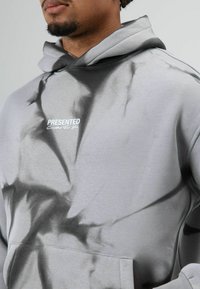 Grijze trui met een rookachtig zwart patroon, voorzien van een voorzak en een capuchon. Tekst "PRESENTED" in het wit op de borst.