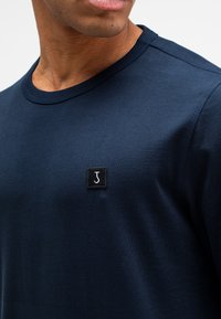 Navyblaue Baumwoll-T-Shirt mit rundem Halsausschnitt und kurzen Ärmeln. Besitzt ein schwarzes Quadrat-Patch mit einem stilisierten weißen "J"-Logo auf der Brust.