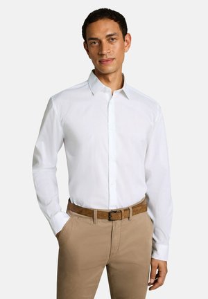 Man die een wit lang mouwen overhemd draagt, stop het in beige broek met een bruine riem, staand met één hand in de zak.