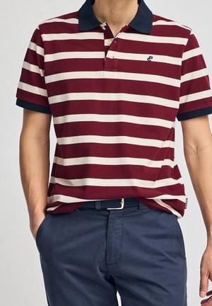 Uomo che indossa una polo a righe bordeaux e bianca con colletto e polsini blu navy, cintura blu navy e pantaloni blu navy con le mani nelle tasche.