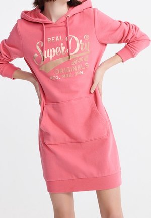 Femme portant une robe sweat à capuche rose SuperDry avec poche avant et texte doré de la marque sur la poitrine.