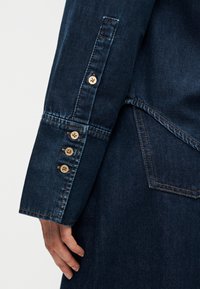 Μανίκι πουκαμίσου από σκούρο τζιν με πέντε ταμπά κουμπιά, ραφές σε αντίθεση και εφαρμογή προσαρμοσμένη στο σώμα, με ορατή λεπτομέρεια τσέπης στο πλάι.