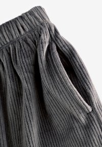 Gros plan sur un pantalon en tissu côtelé gris foncé avec une ceinture élastique et une ouverture de poche latérale visible sur fond blanc.