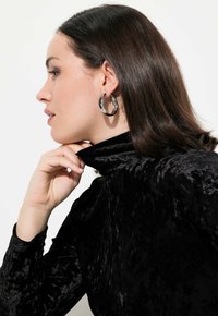 Dolcevita in velluto nero con una texture liscia, caratterizzato da un colletto aderente. Un grande orecchino in argento a cerchio mette in risalto il profilo della modella.