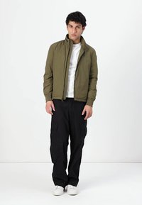 Veste légère vert olive avec fermeture éclair, col montant, portée avec un pantalon cargo noir et des baskets blanches. T-shirt blanc uni en dessous.