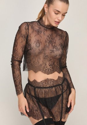 Vrouw draagt een zwarte doorschijnende kanten crop top met lange mouwen en bijpassende doorschijnende kanten rok met jarretels en zwart ondergoed.