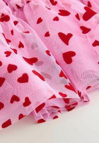 Tissu en tulle rose orné de petits cœurs rouges en feutre, avec une texture douce et des volants superposés, créant une apparence légère et aérienne.