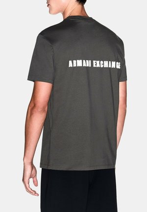 Persona con una camiseta gris oscuro de manga corta con "ARMANI EXCHANGE" impreso en blanco en la parte superior de la espalda, de pie frente a un fondo liso.