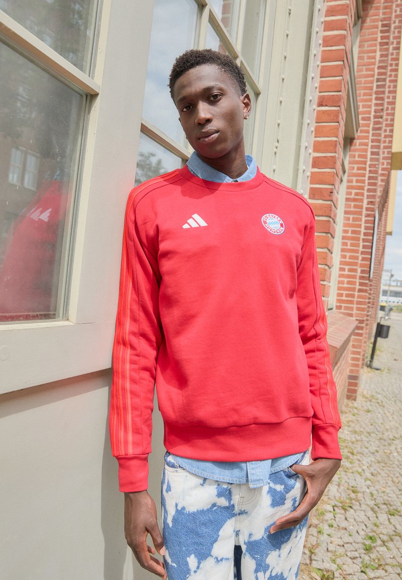 Rød Adidas hettegenser med tre hvite striper på ermene og logo på brystet. Brukt over en lys blå denimskjorte, kombinert med blå tie-dye jeans.