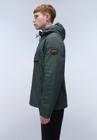 Grøn hættejakke med forlommer og sidelukning, med en rund logo-patch på venstre ærme. Glat tekstur og afslappet design.