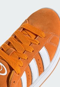 Narancssárga velúr sneaker fehér bőrcsíkokkal, texturált narancssárga fűzőkkel és fehér gumitalppal. Kiemelésként varrási részletek találhatók.