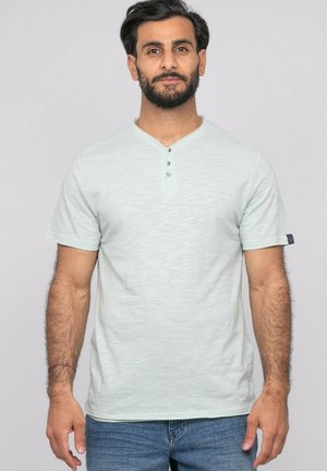Homme barbu portant un t-shirt henley gris clair à manches courtes et un jean bleu, debout devant un fond clair uni.