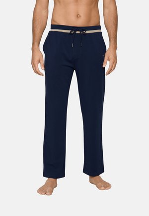 Mann trägt marineblaue Freizeithose mit beigem und marineblauem Taillenband mit Kordelzug, steht barfuß vor einem einfarbigen Hintergrund.