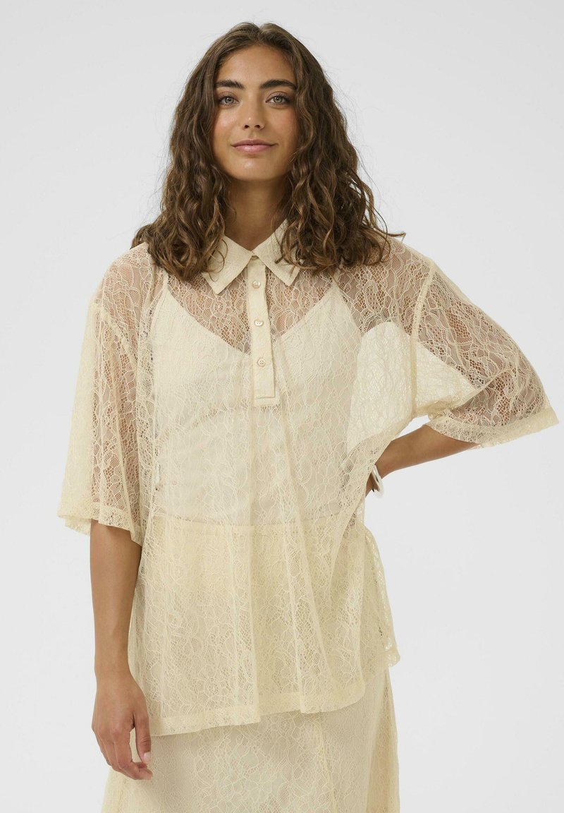 Femme aux cheveux bruns ondulés portant un blouse en dentelle crème avec des boutons et un col, se tenant avec une main sur la hanche devant un fond uni.