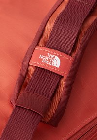 Red and burgundy backpack strap with white "The North Face" logo on a stitched fabric label.