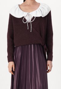 Burgundy stickad tröja med en vit volangkrage och snörning, tillsammans med en plisserad, glänsande lila kjol. Texturerad tyg genom hela plagget.