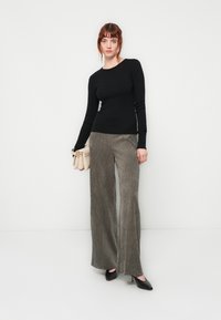 Woman wearing a black long-sleeve top and wide-legged gray corduroy pants, holding a beige handbag, standing against a white background.