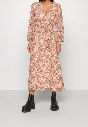 Frau trägt ein langärmeliges, V-Ausschnitt, tailliertes Midikleid mit braunem Leoparden- und pinkem Blumenmuster, kombiniert mit schwarzen klobigen Stiefeletten.