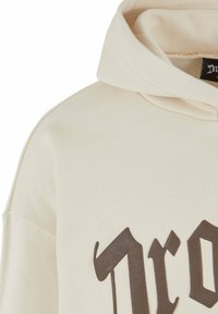 Cream-colored hoodie with a soft texture, featuring a dark brown graphic print and a large, relaxed hood. Simple design with no embellishments.