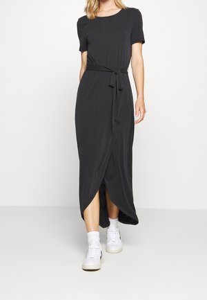 Woman wearing a black, short-sleeve, belted maxi dress with a front overlap slit, paired with white sneakers and socks, walking forward.