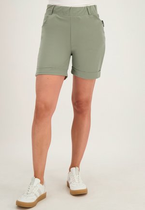 Persoon met lichtgroene shorts met opgerolde zoom en witte sneakers, staand tegen een neutrale achtergrond.
