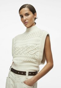 Crèmekleurige kabelgebreide mouwloze sweater met een hoge geribbelde kraag, gekenmerkt door ingewikkelde textuurpatronen, gedragen over lichte broek met een bruine riem.