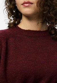 Burgundska pletena jopica z ribasto teksturo, ki ima okroglo ovratnik in raglan rokave, s prikazom mehkega, heather vzorca.